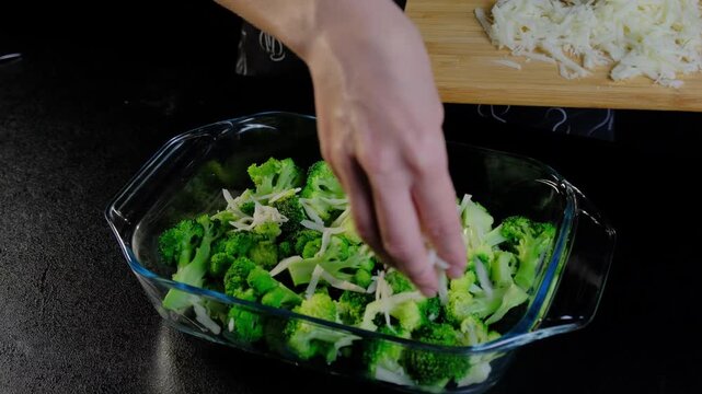Hand sprinkling shredded cheese over broccoli florets in a glass baking dish on a kitchen counter at Cooking.