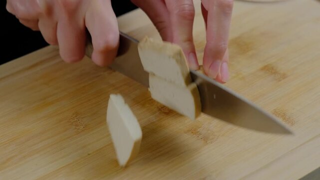 Close-up of adult hands using a silver knife to slice a beige food block into cubes on a wooden cutting board in a Cooking kitchen during daytime.