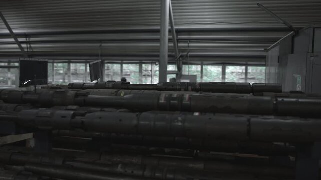 Stacked steel drill rods on horizontal racks inside dim industrial workshop, showing layered metal surfaces, support poles, corrugated ceiling, worn textures, and utilitarian storage