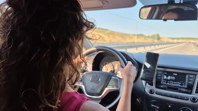 Young woman driving car along highway during sunset, view from back seat. Handheld camera motion follows driver navigating open road during golden hour.