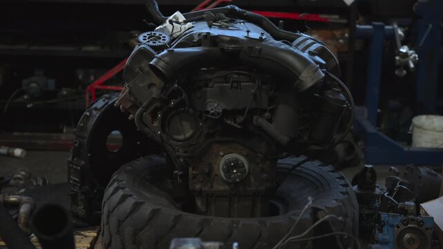 Used internal combustion engine resting on stacked heavy duty tires inside dim workshop, showing intake manifold, valve covers, hoses, pulley, gears, rusted exhaust parts, metal texture