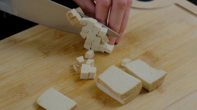 Close-up of hands using a chef's knife to slice tofu on a wooden cutting board in Cooking.