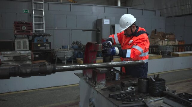 Industrial worker tightening heavy bench vise around metal shaft in workshop, wearing high visibility safety gear under cool overhead lighting beside worn workbench