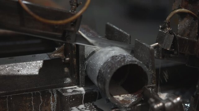 Industrial bandsaw cutting metal pipe with coolant spray in workshop close view, blade engaging clamped cylindrical workpiece with wet guides, sparks, droplets, and machine detail