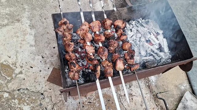 Top view of shashlik skewers on a metal barbecue grill, ash and smoke, rural courtyard with concrete floor, outdoor cooking in a village backyard