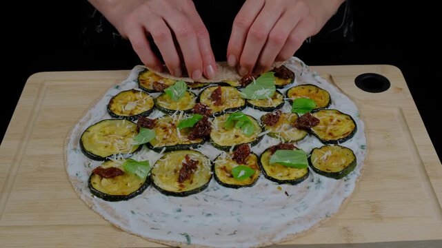 Hands in a kitchen sprinkle breadcrumbs over zucchini slices on a pizza dough base, preparing a savory dish at the Cooking location.