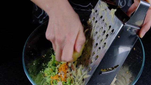 Close-up of hands grating vegetables into a glass bowl in a kitchen, cooking preparation with carrots and greens, daytime.
