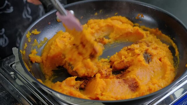 Hands stir bright orange squash puree with spices in a dark frying pan during cooking, using wooden spatula and whisk in a kitchen at Cooking location.