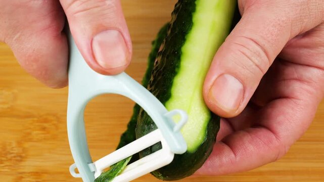 A professional chef uses a white peeler to remove the green skin from a fresh cucumber on a wooden cutting board, exposing pale juicy flesh for healthy gourmet meals.