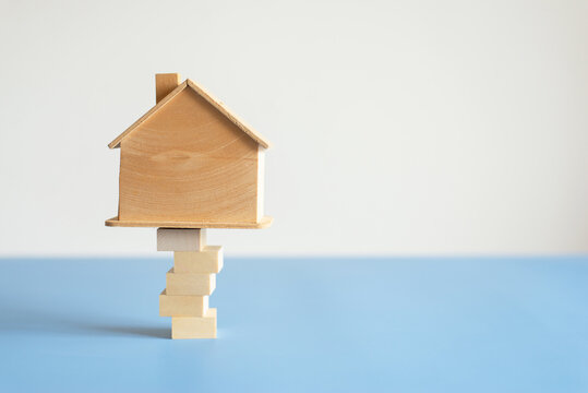 Small wooden house model balancing on unsteady stacked wooden blocks on blue and white background, financial risk and property stability concept