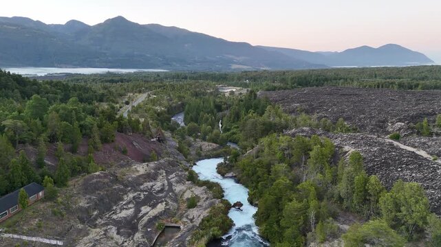 Serene overhead perspective of the bright turquoise Truful Truful River winding through dense coniferous forest interspersed with dark volcanic lava rock fields. Snowcapped Llaima volcano