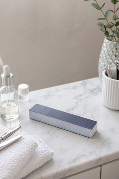 Gray nail buffer block and manicure tools on a white marble table. Cuticle oil and metal instruments prepared for a nail care routine