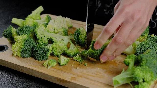Hands in dark clothing chop fresh broccoli florets with a silver knife on a wooden cutting board during food preparation in Cooking.