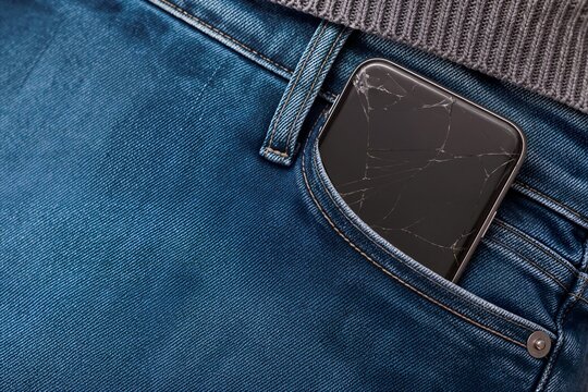 Smartphone with a cracked screen in a denim jeans pocket. Broken mobile device sticking out of blue pants. Technology damage and repair concept