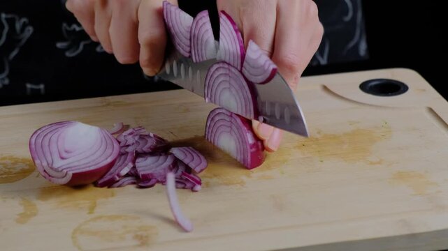 Hands slice red onions on wooden board with knife in a cooking kitchen, bright light, dark background.