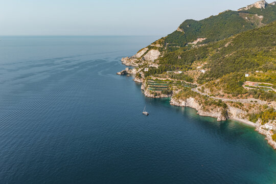 Aerial view of the rugged Amalfi Coast coastline with lush green hills, a winding coastal road, and a small boat floating on the deep blue Tyrrhenian Sea in Cetara, Amalfi Coast, Italy.