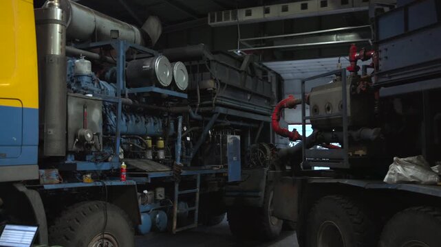Industrial truck mounted diesel engine units inside dim workshop with linked piping, service gap, heavy metal housings, hoses, valves, rust, dust, and maintenance access points
