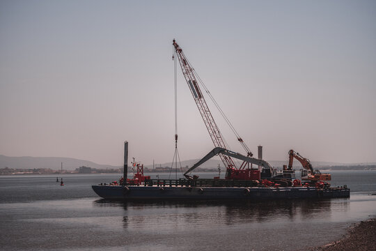 Industrial work barge equipped with a tall lattice boom crane and tracked excavators floating on calm harbor water, with shoreline infrastructure and hazy hills beyond; telephoto shot