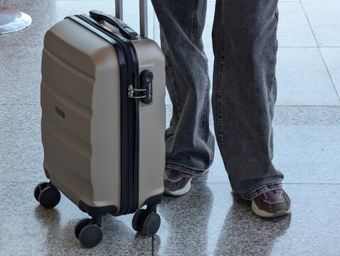 Person with suitcase standing still, in airport terminal, with suitcase, jeans, shoes, from a close-up perspective, with beige, black accents, and neutral mood.