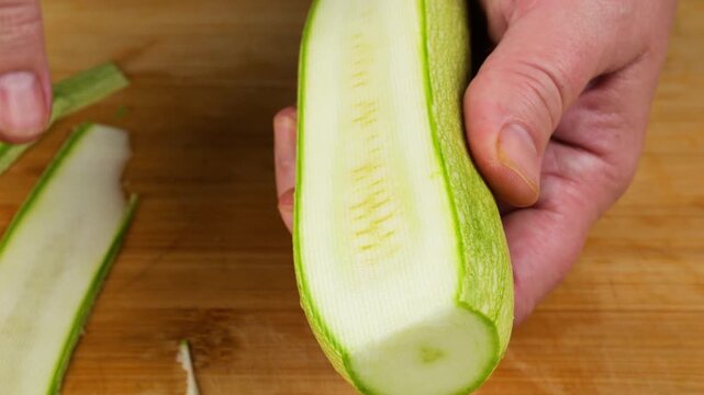 A close-up shot captures a chef using a white vegetable peeler to slice a fresh green zucchini into long, thin translucent ribbons on a wooden board for cooking.
