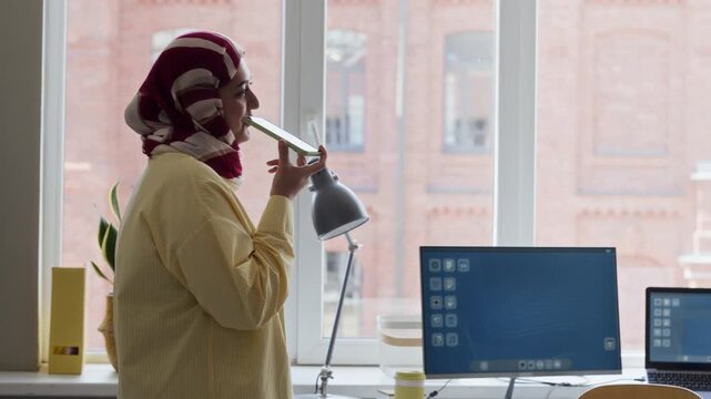 Young adult Muslim woman wearing hijab standing in modern office, recording voice message on smartphone. Clip is ideal for corporate diversity, modern workplace, and mobile technology marketing