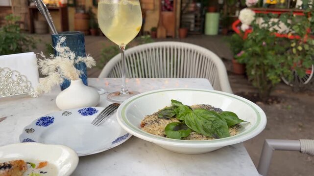 Creamy orzo pasta with sun-dried tomatoes, spinach and caramelized onion served with fresh basil. Italian-style restaurant meal on an outdoor caf&eacute; table with a glass of white wine.