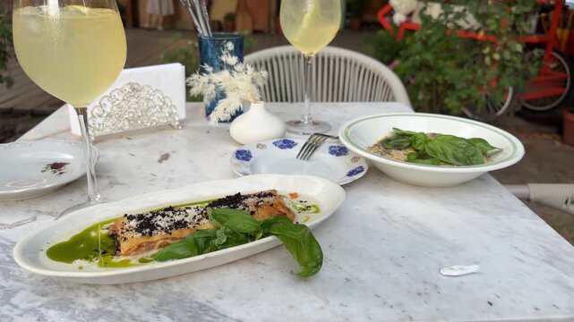 Italian dinner with lasagna, creamy risoni, white wine and fresh basil served on an outdoor restaurant table. Mediterranean cuisine, elegant dining setting, food photography.