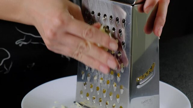 Hands in dark patterned clothing grate yellow cheese on a stainless steel box grater over a white plate in a Cooking kitchen, daytime.