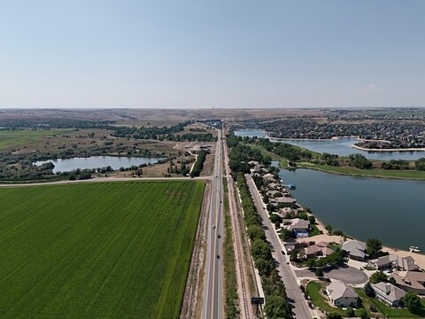 Aerial view of Pelican Lakes Point featuring residential homes along the lakes, green agricultural fields, and a straight road stretching into the horizon in Windsor, Colorado, United States.