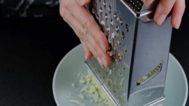 Close-up of hands grating a yellow block of cheese with a metal box grater into a mint green bowl on a dark kitchen countertop during daytime cooking.