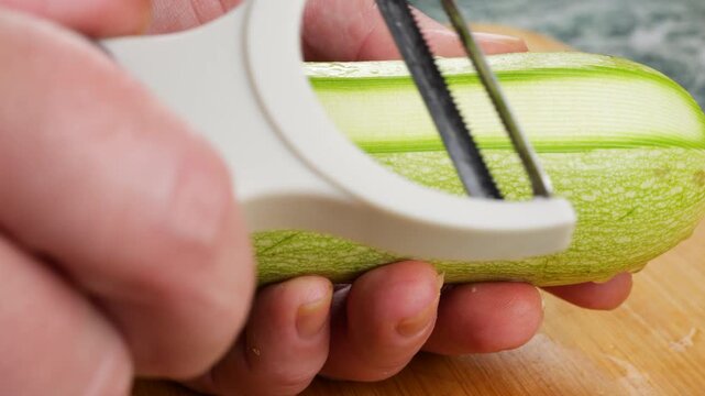 A close-up shot captures a chef using a white vegetable peeler with a sharp stainless steel blade to smoothly remove thin strips of skin from a fresh green zucchini on a wooden board.