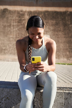 Vertical. Smiling Black Latina athlete using her smartphone while sitting outdoors during a workout break, with a reusable water bottle beside her in bright sunlight.