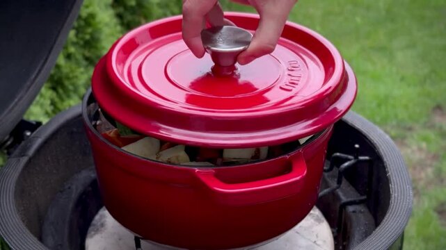 Red clay pot on a barbecue contains a healthy dish of chopped carrots, chunks of meat, and potatoes. The pot is situated on a grill, set within a ceramic barbecue smoker