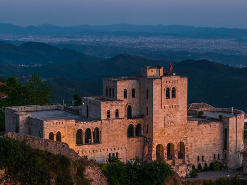 Aerial view of the historic Kruje Castle illuminated by golden hour light, featuring stone walls and the Albanian flag, set against a mountainous landscape in Kruje, Durres County, Albania.
