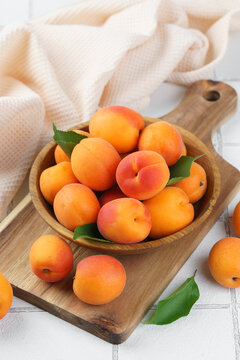 Top view of ripe apricots with green leaves arranged in wooden bowl on on white tile background. Fresh seasonal harvest, summer fruit, healthy eating, dessert, food ingredient