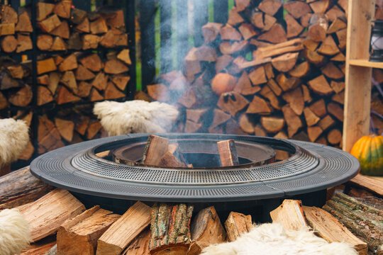 Modern outdoor fire pit, fireplace with smoke surrounded by stacked firewood, soft blankets and autumn decorations. Cozy fall setting for Halloween, Thanksgiving and seasonal outdoor gatherings