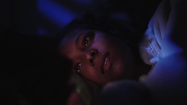 Cheerful African American young woman using smart phone while lying on sofa in dark room at home in the night, illuminated by the cellphone light. Copy space.