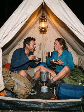 A happy couple enjoys a meal together inside a cozy illuminated camping tent during an outdoor wilderness adventure.