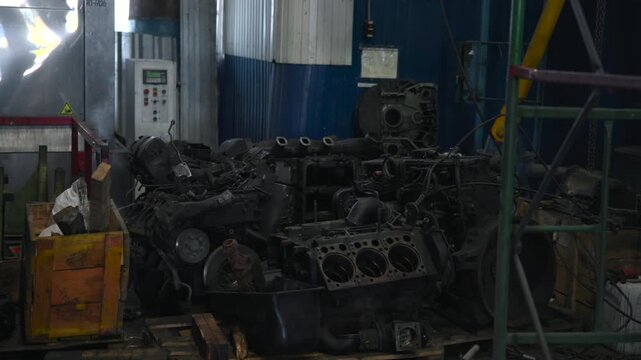 Cluttered industrial workshop interior with stacked disassembled engine components, housings, and pallets under cool diffused light and heavy metal textures, with engine, workshop, machinery, metal