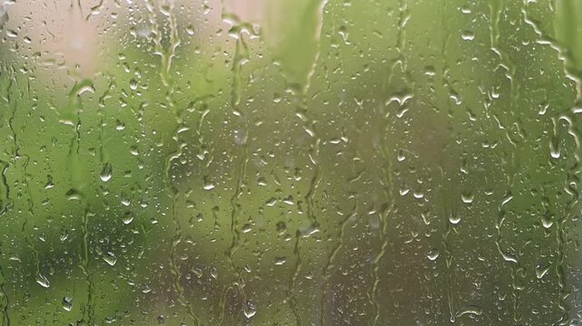 Raindrops falling down a window pane during a storm Heavy rain on glass surface with blurry green nature background