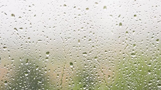 Raindrops falling down a window pane during a storm Heavy rain on glass surface with green nature background blurry