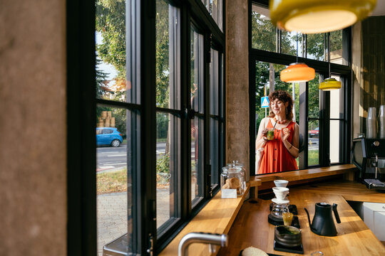 Stylish middle aged woman with iced matcha seen through open cafe window in modern interior