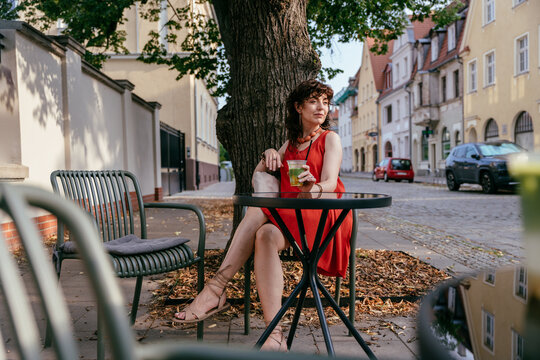 Elegant middle aged woman in orange dress with iced matcha seated at street cafe table