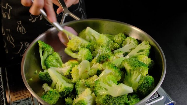 Hands in dark clothing stir broccoli florets in a metal frying pan with a pink spatula on a stovetop in a kitchen, Cooking location.
