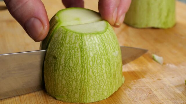 A close-up of a chef using a sharp knife to slice a fresh green zucchini into pieces on a wooden board, preparing healthy organic vegetables for a home-cooked meal.