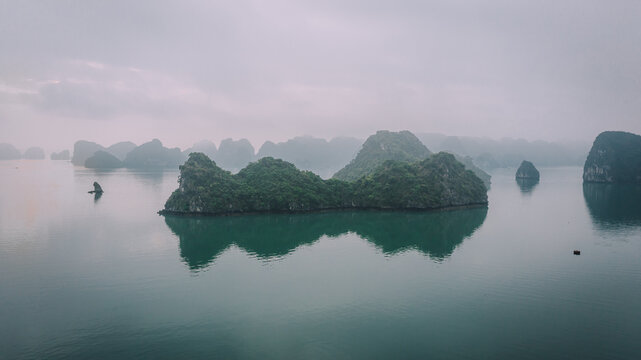Aerial view of Ha Long Bay with limestone karsts rising from the calm, misty waters under an overcast sky in Ha Tu, Quang Ninh, Vietnam.