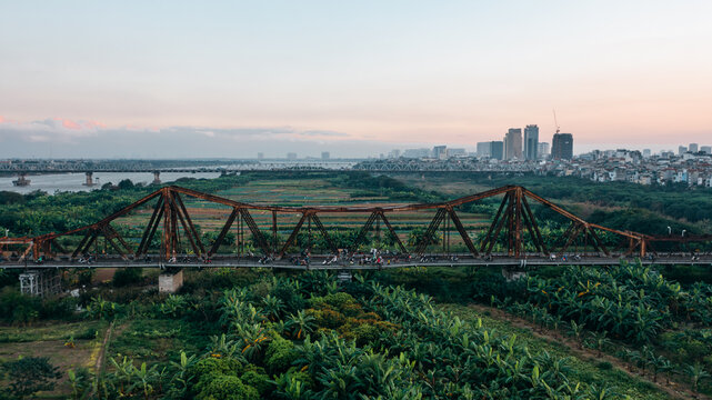 Aerial view of Long Bien Bridge with motorbikes crossing the historic steel structure over the Red River, surrounded by lush vegetation and the Hanoi city skyline in Hong Ha, Hanoi, Vietnam.