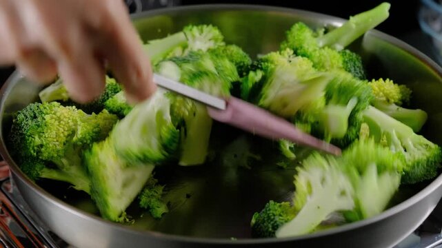 Chef stir-fries chopped broccoli florets in a large metal pan on a stovetop in a kitchen, using a pink spatula, during daytime cooking.