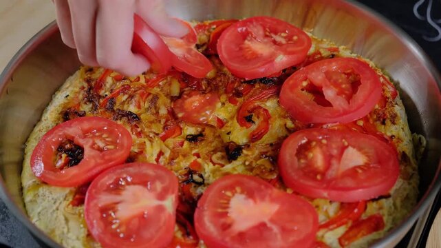 Adult hands arrange red tomato slices on a frittata base with bell peppers in a metal pan, cooking in a kitchen.