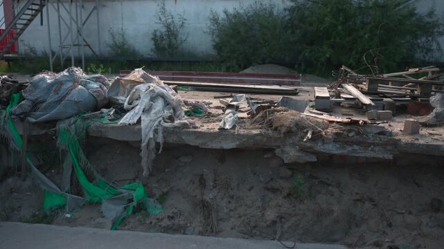 Eroded concrete slab strewn with construction debris in abandoned industrial lot, rough broken edge, exposed soil, scattered rusted metal, weathered wood, frayed fabric, overgrowth, muted sunlight